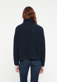 Pull en maille épaisse bleu marine avec un col montant, des épaules tombantes et des motifs en relief en torsades sur les manches. Longueur cropped et coupe décontractée.