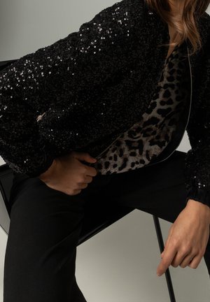 Person iført en sort paljetjakke over en leopardprint top, siddende på en sort stol med den ene hånd, der justerer jakke lynlåsen.