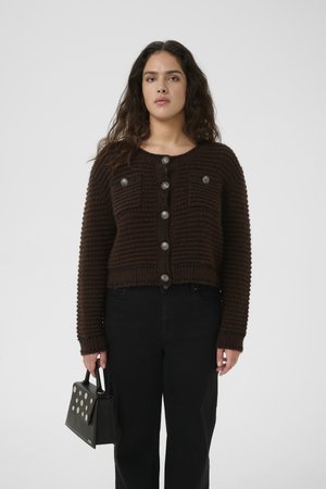 Brown knitted cardigan with a textured pattern, featuring two chest pockets, silver buttons, and a cropped fit, paired with black pants and handbag.