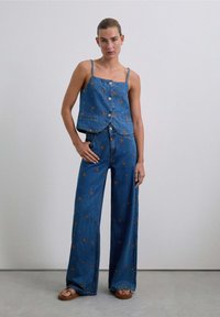 Denim crop top met knopen en spaghetti bandjes, gecombineerd met wijde jeans met bloemembroideries in oranje en blauw op een blauwe achtergrond.