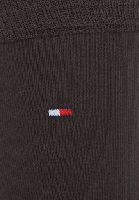 Close-up of dark brown ribbed fabric with small rectangular knit logo in white, red, and navy colors centered on the material.