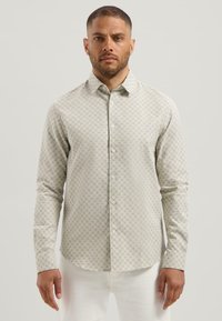 Man in een lange mouwen overhemd met een klein repetitief geometrisch patroon en witte broek, staande tegen een effen achtergrond.