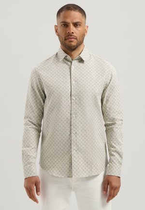 Man in een lange mouwen overhemd met een klein repetitief geometrisch patroon en witte broek, staande tegen een effen achtergrond.