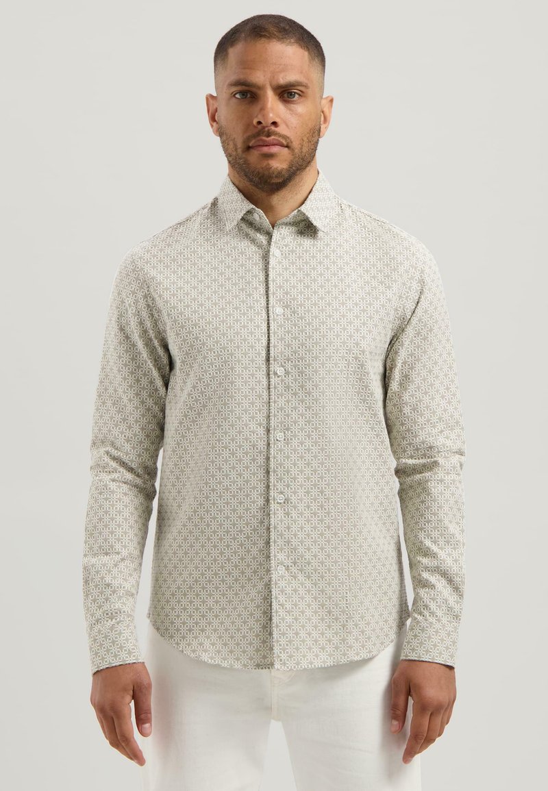 Man in een lange mouwen overhemd met een klein repetitief geometrisch patroon en witte broek, staande tegen een effen achtergrond.