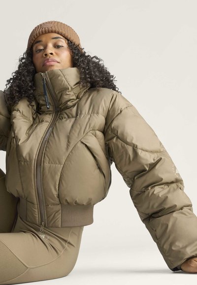 Mujer con pelo rizado que lleva una chaqueta de plumón beige, pantalones a juego y un gorro de punto marrón, sentada frente a un fondo claro.