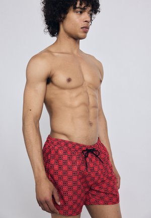 Red swim shorts with a repeating black "GO" pattern. Features an elastic waistband and black drawstring. Smooth fabric, mid-thigh length.