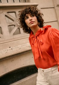 Blouse rouge en soie avec un col noué et des manches longues bouffantes; associée à un pantalon blanc. Le mannequin porte des boucles d'oreilles créoles dorées.