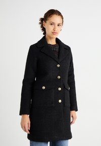 Abrigo de lana negro con un acabado texturizado, que presenta un cuello con muescas, botones dorados, dos bolsillos frontales y un corte entallado.