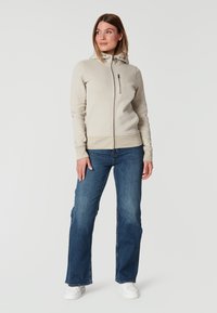 Ljust beige zip-up sweatshirt med hög krage, subtile logodetalj och en framficka. Matcher med blå utsvängda jeans och vita sneakers.
