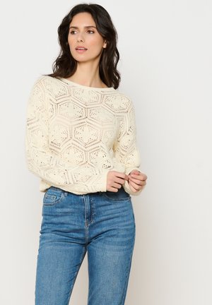Brede crèmekleurige trui van breiwerk met een opengewerkt hexagonaal patroon, lange mouwen en een brede halslijn, gecombineerd met high-waisted blauwe jeans.