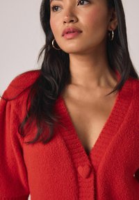 Rdeč pleten pulover z V-izrezom, teksturirano tkanino in gumbom v obliki srca na sredini. Mehki, prijeten videz s kratkimi rokavi.