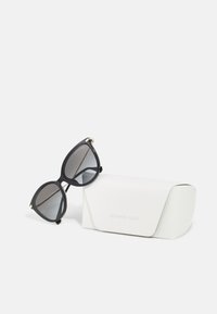 Black oversized sunglasses with gradient lenses, resting on a white textured case. Case features embossed brand name.