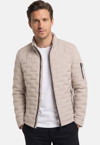 Mann trägt eine beige gesteppte Jacke über einem weißen Hemd, mit den Händen in den Taschen, vor einem schlichten hellen Hintergrund.