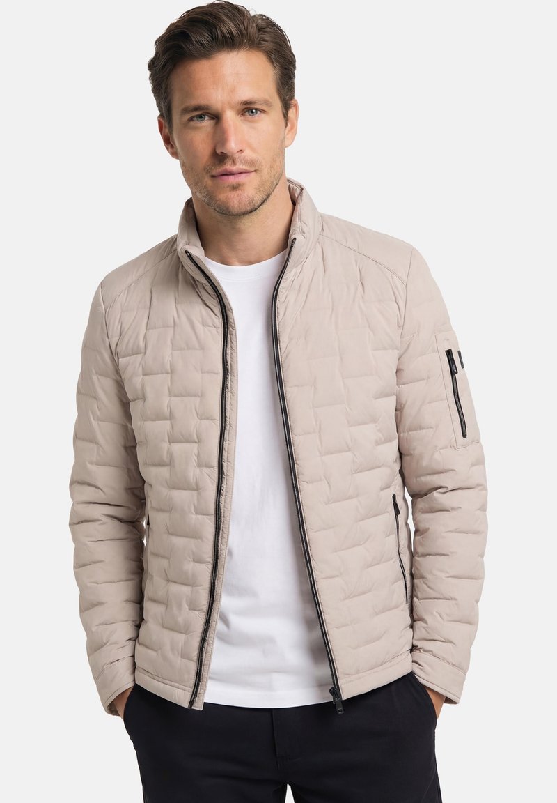 Mann trägt eine beige gesteppte Jacke über einem weißen Hemd, mit den Händen in den Taschen, vor einem schlichten hellen Hintergrund.