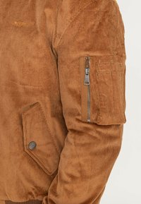 Velours bomberjack in warm bruin met geribbelde textuur, voorzien van een ritszak op de mouw, drukknoop en geborduurd detail op de borst.