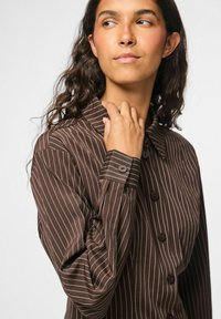 Brun stribet skjorte lavet af stof med krave, med knaplukning og lange ærmer. Striberne er i en lysere nuance på en mørk baggrund.