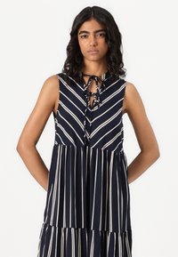 Vestido sem mangas azul-marinho com riscas brancas verticais, apresentando um decote atado e um design de saia em camadas. Textura de tecido suave.