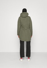 Olivgrön, lång, vadderad jacka med huva, åtdragen midja och sidofickor, bärs över svarta byxor och vita sneakers med röda detaljer.