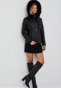 Giacca in pelle nera con cappuccio bordato di pelliccia, con due tasche sul petto e un fit sartoriale. Abbinata a una gonna nera texturizzata e stivali al ginocchio.