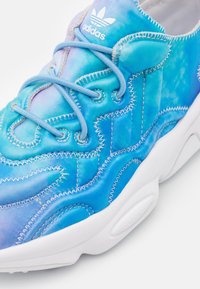 Blue and teal sneaker with a wavy stitched pattern, fabric upper, and white rubber sole. Features rounded laces and an Adidas logo.