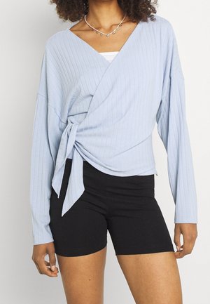 Mujer con un top cruzado azul claro acanalado atado a la cintura y pantalones cortos negros ajustados con un collar plateado.