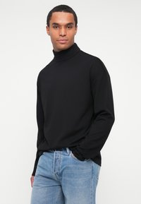 Maglione a collo alto nero realizzato in un tessuto liscio, con maniche lunghe e un fit rilassato, abbinato a jeans di denim azzurro chiaro.