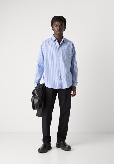Chemise boutonnée rayée bleu et blanc, manches longues, poche poitrine unique, associée à un pantalon noir et des chaussures noires, tenant une veste en cuir.