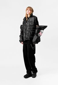 Chaqueta tipo plumón de tela negra brillante con grandes bolsillos, mangas amplias con un diseño de oso de peluche, llevada sobre una-shirt negra y pantalones holgados.