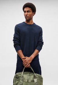 Tumši zils adīts džemperis ar garām piedurknēm, apaļu izgriezumu un zaļu akcentētu šuvju detaļu, kombinēts ar teksturētu olīvu zaļu somiņu.