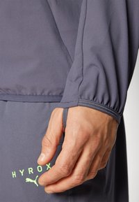 Graues sportliches Langarmshirt mit gerafftem Bündchen, das ein grünes "HYROX"-Logo und einen glatten, leichten Stoff aufweist. Hand in der Nähe der Naht positioniert.