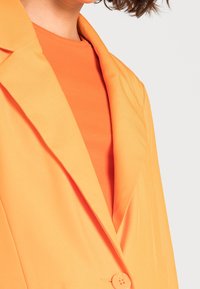 Oranger Blazer mit Reverskragen, taillierter Passform und strukturiertem Innenfutter. Die Knopfdetails sind an der Vorderseite sichtbar.