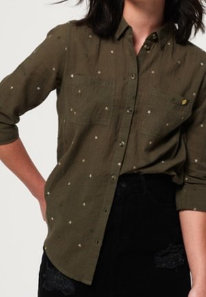 Mujer con una camisa verde oliva abotonada con pequeños patrones de estrellas blancas y pantalones negros con parches de encaje.