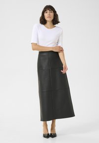 Falda maxi de cuero negro con detalles en panel, combinada con una blusa blanca ajustada de manga corta y zapatos de tacón negro de punta en un fondo liso.