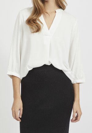 Kobieta ubrana w białą bluzkę z dekoltem w serek i rękawami do połowy ramienia, włożoną do czarnej, prążkowanej, wysokopodniesionej spódnicy, stoi na tle białego tła.