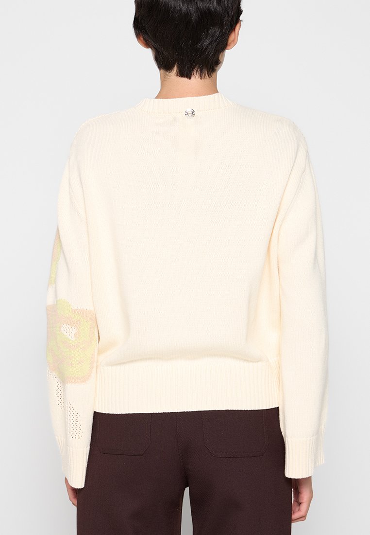Person, der einen cremefarbenen Strickpullover mit geripptem Saum und Bündchen trägt, mit floralen Mustern auf dem linken Ärmel, und dunklen braunen Hosen, von hinten betrachtet.