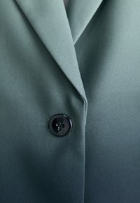 Blazer aus einfarbigem olivgrünem Stoff mit glatter Textur, ausgestattet mit einem scharfen Kragen und einem einzelnen schwarzen Knopfdetail.