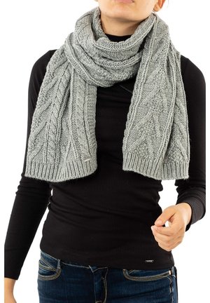 Personne portant une épaisse écharpe grise en tricot avec des motifs torsadés, par-dessus un col roulé noir côtelé et un jean bleu.
