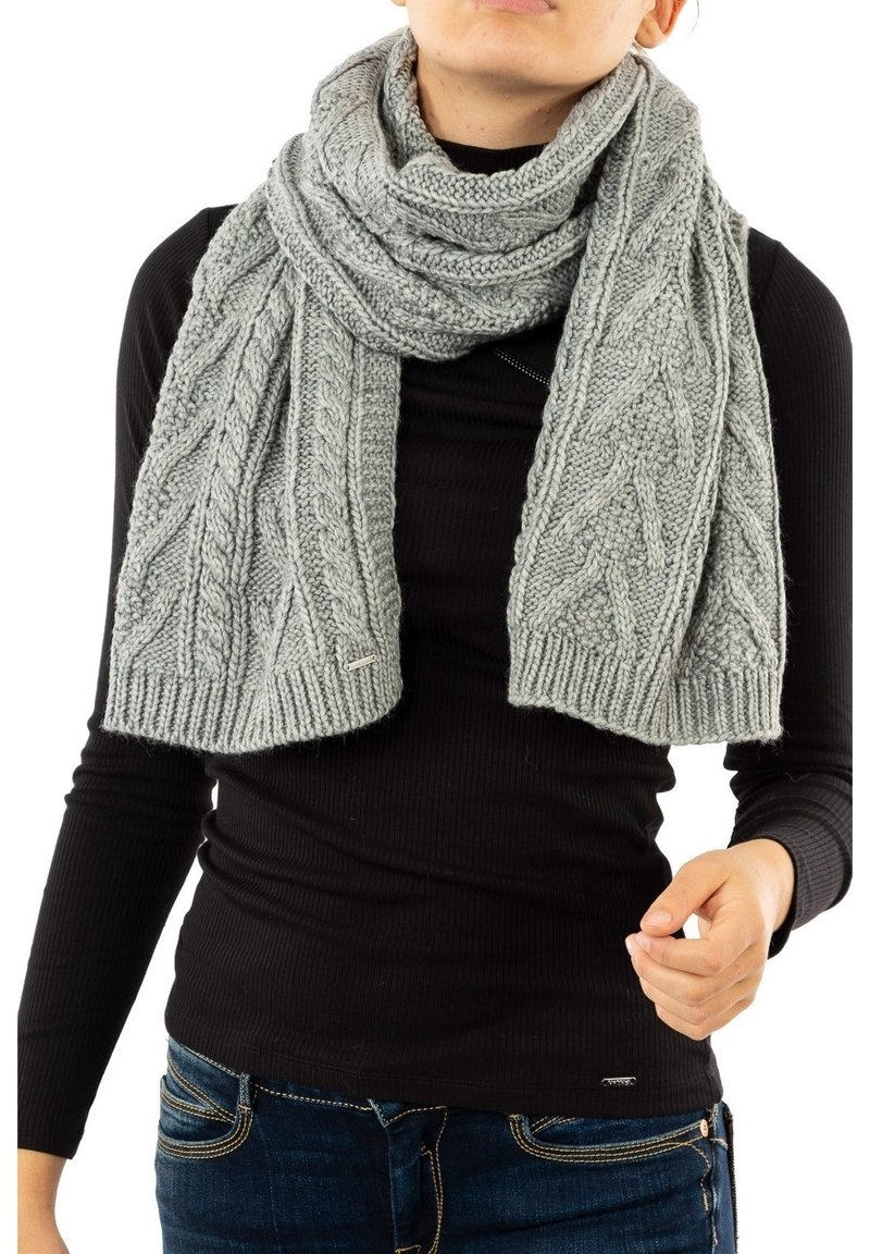 Personne portant une épaisse écharpe grise en tricot avec des motifs torsadés, par-dessus un col roulé noir côtelé et un jean bleu.