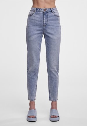 Spodnie jeansowe slim fit z wysokim stanem w jasnoniebieskim kolorze, noszone z jasnoszarymi sandałami na platformie z odkrytymi palcami, na osobie stojącej na białym tle.