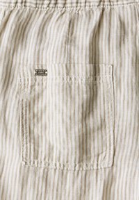 Lichtgekleurde stof met verticale strepen in beige en crème. Bevat een achterzak met stikseldetails en een klein label met de tekst "Three One Studio."