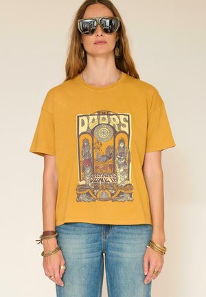 Femme portant un t-shirt jaune moutarde avec un graphisme vintage de The Doors, un jean bleu, des bracelets dorés et de grandes lunettes de soleil réfléchissantes.