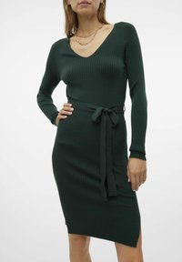 Robe tricotée verte à côtes avec manches longues, décolleté en V, ceinture à la taille et fente latérale. Présente une silhouette ajustée et un tissu texturé.