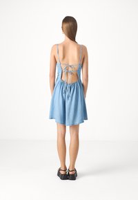 Robe en denim bleu clair avec des bretelles spaghetti réglables, un dos ouvert, une taille froncée et une jupe fluide, associée à des chaussures noires.