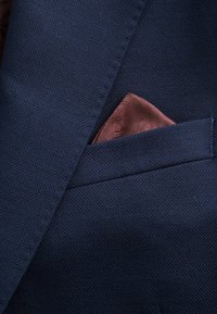 Granatowy materiał garniturowy o teksturze z złożonym jasnobrązowym wzorzonym chusteczką widoczną w kieszeni piersiowej. Garnitur ma precyzyjne detale szycia.