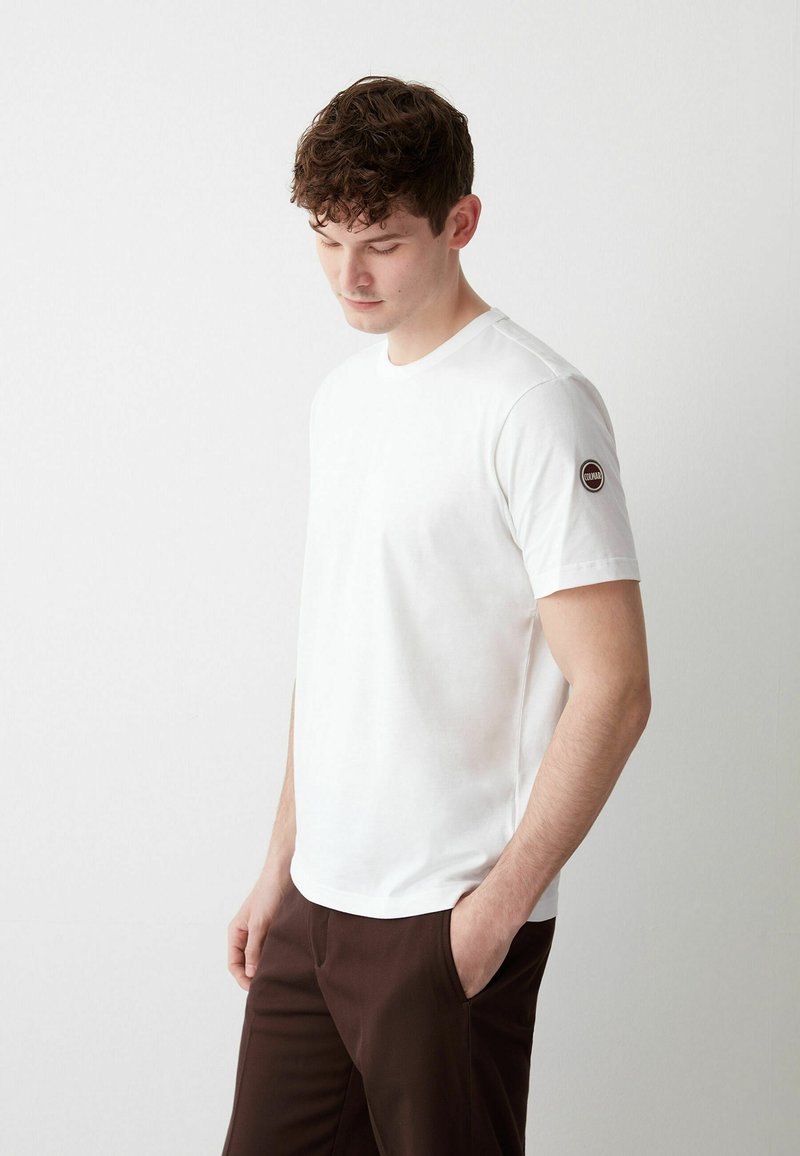 T-shirt bianca in cotone con scollatura rotonda, maniche corte e un patch circolare con logo sulla spalla sinistra, abbinata a pantaloni marroni.