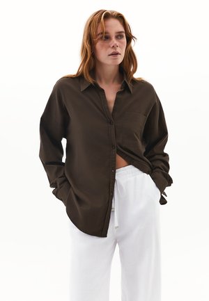 Camicia marrone con bottoni, dotata di taschino sul petto, con una vestibilità comoda e maniche lunghe, abbinata a pantaloni bianchi larghi.
