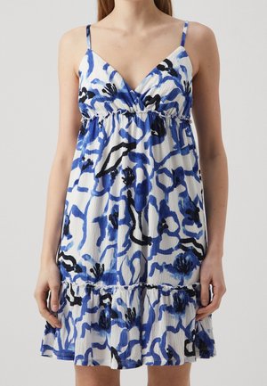 Mujer con vestido blanco sin mangas con estampado floral abstracto en azul y negro y dobladillo con volantes.