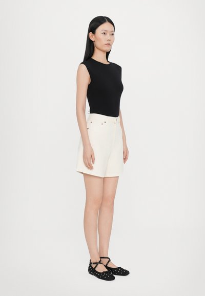 Μαύρο αμάνικο τοπ, λευκά σορτς με ψηλή μέση και μαύρα επίπεδα παπούτσια με πουά. Απλό φόντο, έμφαση στις λεπτομέρειες της ένδυσης.