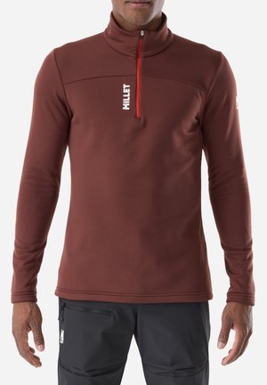 Mann trägt einen braunen Langarm-Pullover mit rotem Reißverschluss und weißem "MILLET"-Logo, kombiniert mit schwarzen Hosen mit Reißverschlusstaschen.