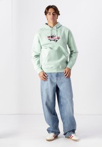Světle zelená mikina s kapucí a grafikou auta na hrudi, přední klokanková kapsa a bílé šňůrky. Kombinováno s volnými modrými džíny a teniskami.